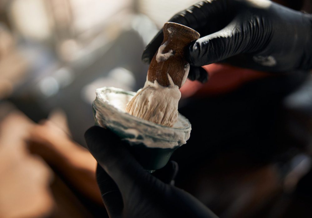 Close up of man hairdresser in sterile gloves putting shave brush into bowl with shaving cream